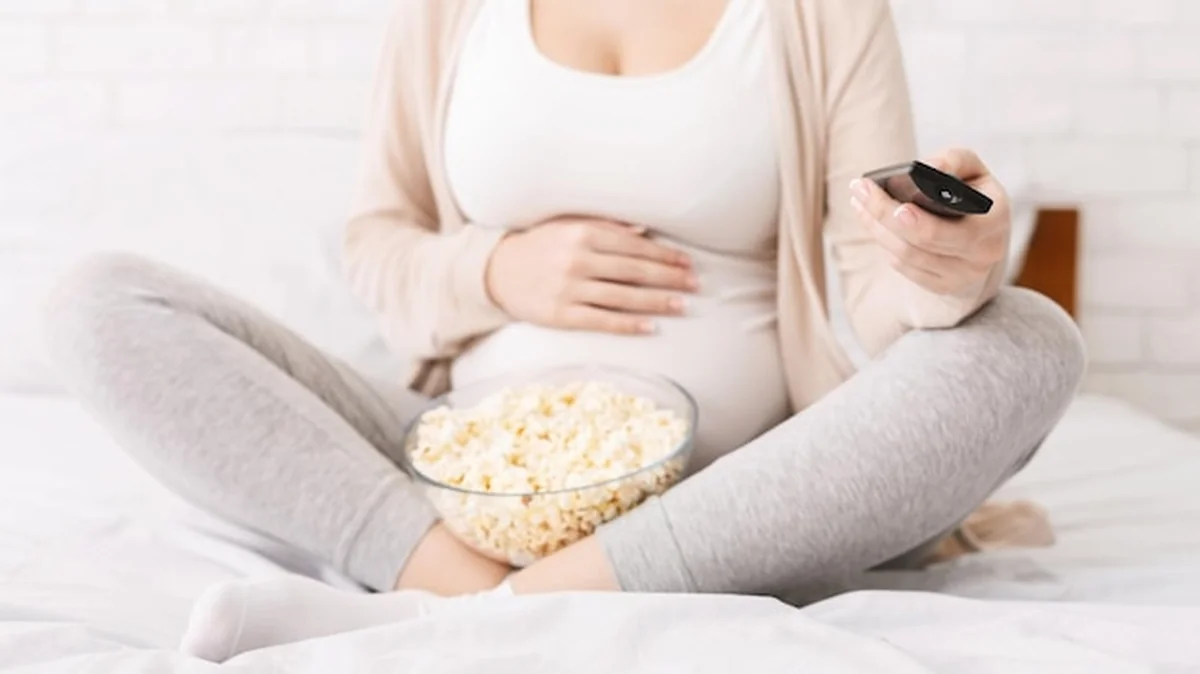 Ciężarna kobieta siedząca na łóżku z miską popcornu i zdalnym sterowaniem