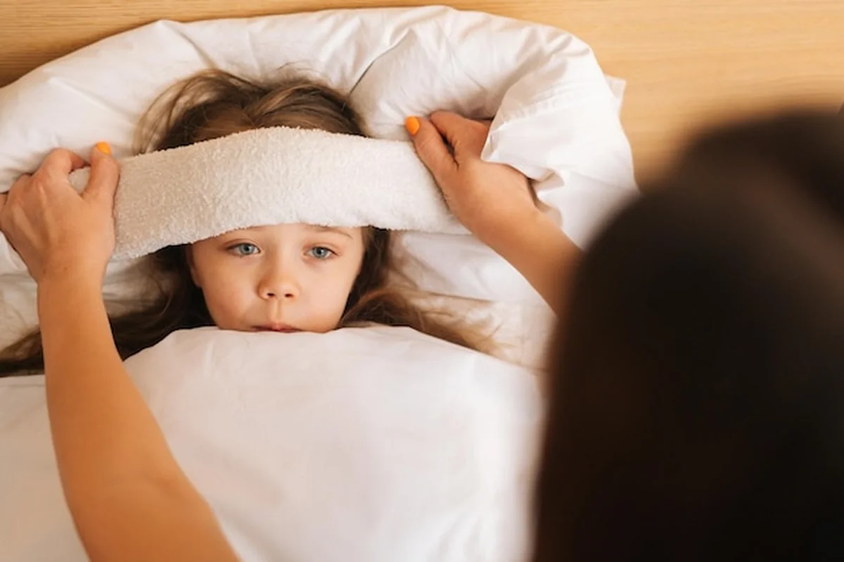 Matka przykłada kompres na czoło chorej córki leżącej pod kocem w domu